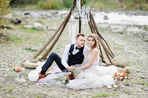 Attractive couple newlyweds, happy and joyful moment. Man and woman in festive clothes sit on the stones near the wedding decoration in boho style. Ceremony outdoors. photo