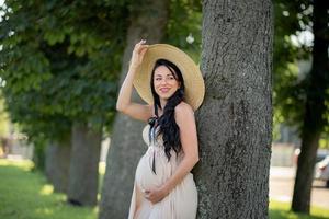 mujer embarazada con un sombrero posando con un vestido sobre un fondo de árboles verdes. foto