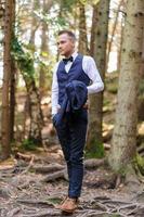 A serious portrait of a handsome groom in a blue suit and bow tie is standing against the backdrop of greenery in the forest. photo