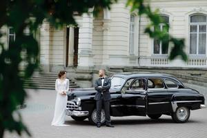 elegante novia hermosa y novio guapo abrazándose en un elegante coche negro a la luz. vista inusual desde atrás. pareja de bodas de lujo en estilo retro. foto