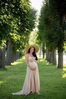Pregnant woman in a hat posing in a dress on a background of green trees.