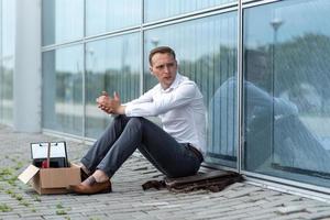 The fired office worker fell to his knees and covered his face due to stress. In front of him is a cardboard box with stationery. The man is unhappy due to a reduction in the robot. photo