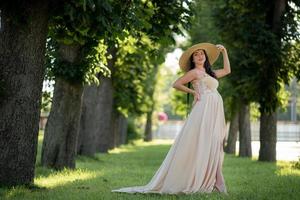 Pregnant woman in a hat posing in a dress on a background of green trees.