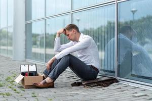 The fired office worker fell to his knees and covered his face due to stress. In front of him is a cardboard box with stationery. The man is unhappy due to a reduction in the robot. photo