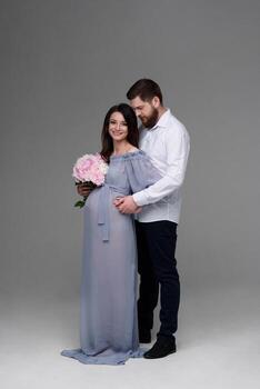 A pregnant woman and her husband are hugging on a gray background in the studio. photo