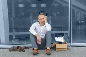 The fired office worker fell to his knees and covered his face due to stress. In front of him is a cardboard box with stationery. The man is unhappy due to a reduction in the robot. photo