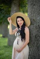 mujer embarazada con un sombrero posando con un vestido sobre un fondo de árboles verdes. foto