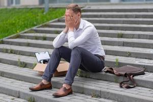 The fired office worker fell to his knees and covered his face due to stress. In front of him is a cardboard box with stationery. The man is unhappy due to a reduction in the robot. photo