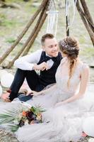Attractive couple newlyweds, happy and joyful moment. Man and woman in festive clothes sit on the stones near the wedding decoration in boho style. Ceremony outdoors. photo