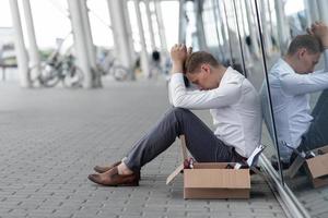 The fired office worker fell to his knees and covered his face due to stress. In front of him is a cardboard box with stationery. The man is unhappy due to a reduction in the robot. photo