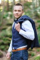 A serious portrait of a handsome groom in a blue suit and bow tie is standing against the backdrop of greenery in the forest. photo