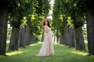 mujer embarazada con un sombrero posando con un vestido sobre un fondo de árboles verdes. foto