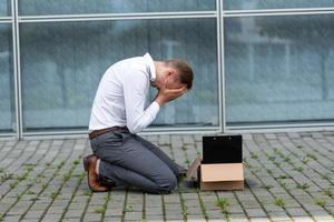 The fired office worker fell to his knees and covered his face due to stress. In front of him is a cardboard box with stationery. The man is unhappy due to a reduction in the robot. photo