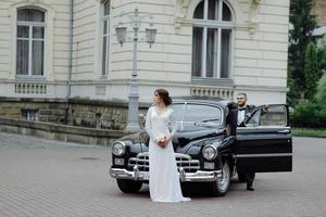 feliz pareja de bodas de lujo besándose y abrazándose cerca de retro con coche de ramo en otoño foto