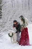 Beautiful bride and groom with a white dog are standing on the background of a snowy forest. photo