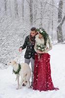 Beautiful bride and groom with a white dog are standing on the background of a snowy forest. photo