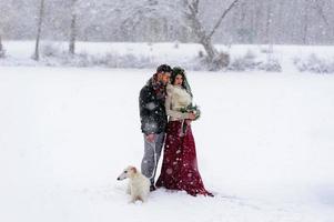 Beautiful bride and groom with a white dog are standing on the background of a snowy forest. photo