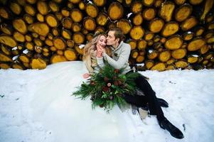 Groom kisses his bride on the temple. Newlyweds with bouquet sits on snow on the wooden background. Winter wedding photo