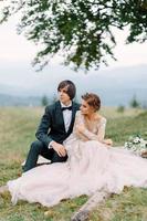 Attractive couple newlyweds, happy and joyful moment. Man and woman in festive clothes sit on the stones near the wedding decoration in boho style. Ceremony outdoors. photo