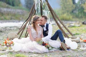 una atractiva pareja de recién casados, un momento feliz y alegre. un hombre y una mujer se afeitan y se besan con ropa de fiesta. ceremonia de boda de estilo bohemio en el bosque al aire libre. foto