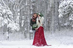 hermosa novia y novio con un perro blanco están parados en el fondo de un bosque nevado. foto