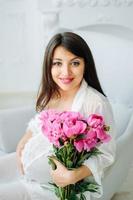 Close up photo of a pregnant woman with a naked belly holding a baby white booties and pink flowers on a white bed