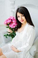 Close up photo of a pregnant woman with a naked belly holding a baby white booties and pink flowers on a white bed