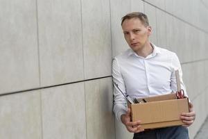 The fired office worker fell to his knees and covered his face due to stress. In front of him is a cardboard box with stationery. The man is unhappy due to a reduction in the robot. photo