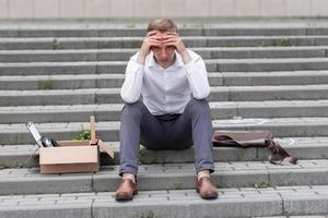 The fired office worker fell to his knees and covered his face due to stress. In front of him is a cardboard box with stationery. The man is unhappy due to a reduction in the robot. photo