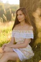 Portrait of a young beautiful girl in a sundress. Summer photo session in the park at sunset. A girl sits under a tree in the shade.