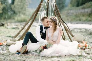 Attractive couple newlyweds, happy and joyful moment. Man and woman in festive clothes sit on the stones near the wedding decoration in boho style. Ceremony outdoors. photo