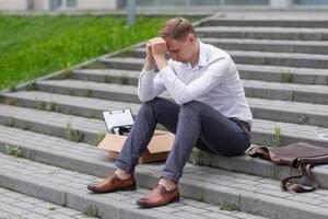 The fired office worker fell to his knees and covered his face due to stress. In front of him is a cardboard box with stationery. The man is unhappy due to a reduction in the robot. photo