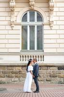 Wedding photo session on the background of the old building. The groom watches his bride posing. Rustic or boho wedding photography.