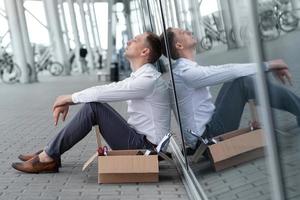 The fired office worker fell to his knees and covered his face due to stress. In front of him is a cardboard box with stationery. The man is unhappy due to a reduction in the robot. photo