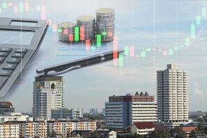 doble exposición de calculadora de negocios, montón de monedas, gráfico financiero y bolígrafo en el escritorio de la oficina, gráfico de acciones, vista de la ciudad en segundo plano foto