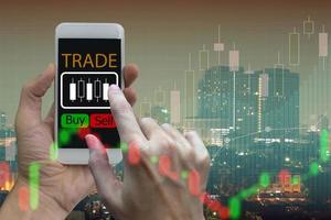 Hand use smartphone trading online on screen with Candle stick graph chart of stock market and city background photo