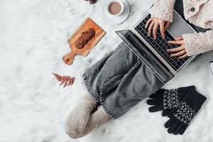 una chica con ropa de invierno que trabaja con una laptop en casa y disfruta de la temporada de invierno con chocolate caliente y galletas foto