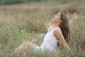 hermosa mujer joven sentada en el campo de hierba verde y soplando diente de león. al aire libre. disfruta de la naturaleza. niña sonriente saludable en el césped de primavera. concepto libre de alergias. libertad foto