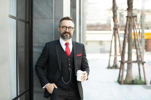 hombre de negocios hipster en traje de pie con una taza de café cerca de la oficina al aire libre foto