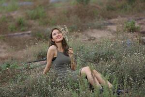 hermosa mujer joven sentada en el campo de hierba verde y soplando diente de león. al aire libre. disfruta de la naturaleza. niña sonriente saludable en el césped de primavera. concepto libre de alergias. libertad foto