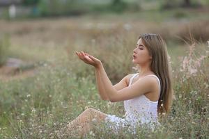 hermosa mujer joven sentada en el campo de hierba verde y soplando diente de león. al aire libre. disfruta de la naturaleza. niña sonriente saludable en el césped de primavera. concepto libre de alergias. libertad foto