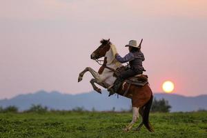 vaquero montando a caballo con pistola de mano, vaquero a caballo contra una hermosa puesta de sol, vaquero y caballo a primera luz, montaña, río y estilo de vida con fondo de luz natural foto
