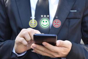 Businessman pressing face emoticon in smartphone at display on virtual screen. Concept of customer service satisfaction evaluation and feedback. photo