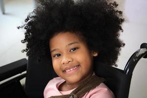 African - American ethnicity little girl resting on wheelchair while waiting to see the doctor in hospital. African - American girl sitting on a wheelchair and smiling to camera. photo