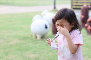 encantadora niña asiática de 4 años, pequeña niña enferma en edad preescolar que sopla la nariz con síntomas de gripe tosiendo al aire libre. copie el espacio concepto de gripe foto