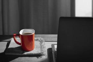 Focus on red coffee cup on napkins with blurred black laptop on wooden table and sunlight shine through the window in living room background,  coffee break concept, splash red color technique photo