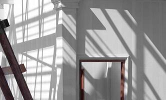 Sunlight and shadow of scaffolding with roof structure on surface of wooden door frame in white primer wall inside of house building site with blurred wooden ladder on foreground, side view with space photo