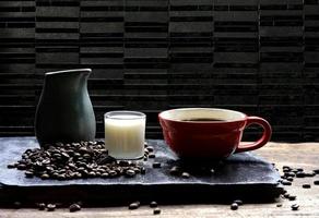 enfoque selectivo en la taza de café roja con leche y granos de café en pizarra negra y mesa de madera en fondo de pared de baldosas de mármol negro foto