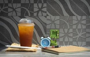 Fresh ice tea in takeaway glass with straw, notebook, pen, blue alarm clock and little wooden picture frame on the table with tile wall decoration background, refreshing in leisure activity concept photo