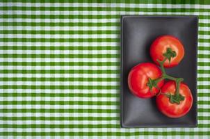 Flat lay of fresh organic tomatoes in black plate on green plaid tablecloth photo